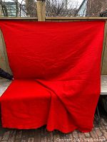 Photo showing large red wool blanket draped and partially folded on a bench outdoors, bright solid red color, visible creases and folds.