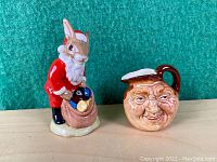 Bunnykins figurine dressed as Santa next to smiling face Toby jug on wooden surface with green background.