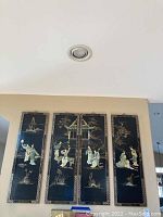 Full view of four black decorative panels with hand-painted Asian style figures and natural elements, mounted on a wall.