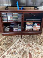 Wooden entertainment cabinet with multiple shelves showing stacks of CDs in jewel cases and DVDs in plastic cases along with the LG Blu-Ray player