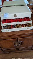 White jewelry chest partly open showing upper compartment and two drawers filled with costume jewelry