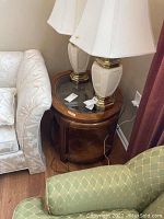 Wooden glass top end table positioned between a white upholstered sofa and a green patterned armchair, with two ceramic lamps on top.
