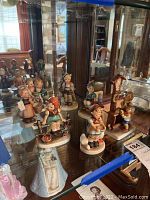 Seven Goebel Hummel porcelain figurines displayed inside a glass cabinet showing various children in traditional clothing and poses including standing, sitting, and interacting near a wooden clock structure.