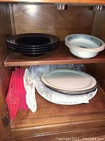 Shelves in wooden cabinet holding six black bowls and two Lenox Autumn patterned bowls.