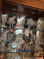 Full shelf view showing multiple crystal stemware pieces and Mikasa plates on display in wood and glass cabinet.