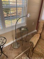 Photo of wrought iron plant stand and Lamicall adjustable phone holder near window by chair.