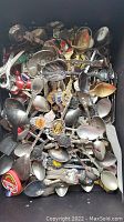 Full box of mixed souvenir spoons piled together showing various spoons with colorful enamel and metal finishes.