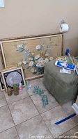 View of green cube storage stool with personal care items on top, four green foo dog figurines on floor, and framed floral and circular art leaning against wall