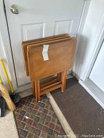 Four wooden tray tables stacked together with foldable legs, showing wood grain and surface wear.