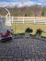 Overview of outdoor planters including two beige pedestal planters with faux flowers and a black rectangular planter with faux red flowers, located on stone patio with grassy yard in background.