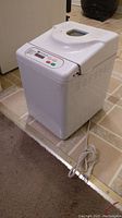 Closed view of the white bread maker showing power cord and top control panel with buttons and display.