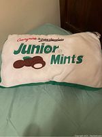 Front view of white pillow with green trim displaying Junior Mints logo and chocolate mint illustration.