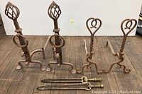 Full view of four wrought iron andirons with two brass fireplace tongs on wooden floor.
