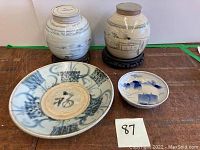 Photo showing two antique ginger jars with black painted Asian landscapes on top of carved wooden stands, one large ceramic plate with blue abstract patterns, and a small ceramic bowl with blue designs.