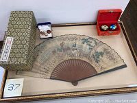Wide view of entire lot showing framed Chinese painted paper fan, cloisonné cufflinks in blue box, decorative vase in box, and metal balls in red box