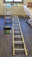 Full view of two aluminum extension ladders, one step stool, Dewalt drill case, and assorted drill bits on wooden floor near workbench.