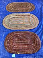 Three oval braided rugs laid out on blue tarp; top small brown-toned, middle medium pastel-toned, bottom large red-toned rug
