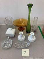 Full view of all vintage glassware items including green vase, amber pedestal bowl, clear pitcher, milk glass covered dish, advertising bottle, two white candlestick holders, and two cut crystal dishes.