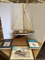 Model sailboat displayed on a wooden stand with two nautical books and a signed painting arranged at its base.