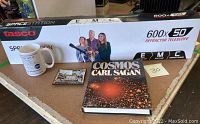 Photo showing the boxed Tasco Spacestation telescope with 600x50 refractor specifications, white NASA mug with logo, Apollo commemorative tray, and Carl Sagan Cosmos book on table.