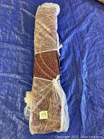Rolled vintage braided rug wrapped in plastic sheeting and tied with a white cord, with a tag marked '89'