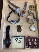 Photo showing various men's wristwatches, cufflinks, pins, and small jewelry items arranged inside a shallow wooden box.