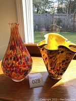 Two Paradigm art glass vases on wooden table by window, showing the orange tall vase and yellow ruffled vase.