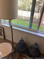 Photo showing brass floor lamp with white cylindrical shade, bentwood and cane accent table, and two decorative ceramic pieces on carpet