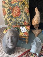 Group photo showing three face sculptures placed on and in front of a floral patterned footstool with golden legs and a pillow upholstery.