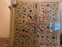 Full view of the framed embroidered textile art showing its square shape and colorful floral motifs arranged in a central circular pattern and surrounding grid.