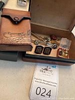 Photo of Berluti leather box open showing men's accessories including cufflinks, tie bars, and tie tacks inside.