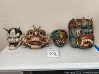 Four masks arranged on a white surface including two large wooden Khon style masks with horns and fangs and two smaller ceramic masks with detailed features.