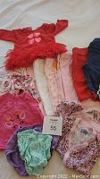 Photo of assorted baby girl clothing including a red dress with heart designs, various onesies, pants, and bloomers