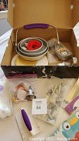 Box containing round metal pans including a red circular cutter, a white piping tool with purple handle, piping bags, and various decorating tips.
