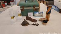 Table display with two white glass oil lamps, metal door man bell, wooden shoe last, vintage eyeglasses, boxed miniature men's shoes, cylindrical Kodak roll container, matchbox, paper ephemera, and vintage green train case.