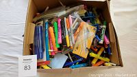 View capturing the wide assortment of various pens and markers in a cardboard box, showing the mixed condition and quantity of items.
