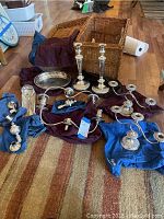 Full lot view showing the four silver tone candelabras, serving dish and spoon, all arranged around the wicker basket on a hardwood floor.