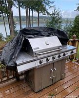 Jenn-Air stainless steel gas BBQ grill with black protective cover, showing control knobs and outdoor setting.