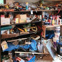 Wide view of garage shelving filled with assorted DIY supplies including paint cans, auto fluids, boxes, and various tools scattered and stacked on shelves and the floor.