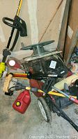 Overall photo showing Toro lawn mower, Ryobi weed wacker, hedge trimmer, red fuel container, and green hose wheel in a storage garage corner
