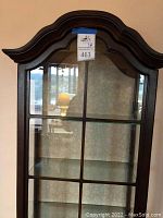 Full frontal view of the wood and glass cabinet showing the decorative curved top, paneled glass door, and drawers at the base.