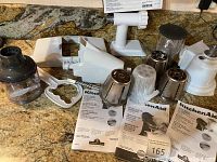 Full spread of KitchenAid mixer attachments on granite countertop including slicer/shredder drums, sausage stuffer, grinder parts, and manuals.