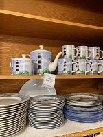 Shelf with lighthouse-themed teapot, creamer, sugar bowl, and coffee mugs above stacks of plates with lighthouse design
