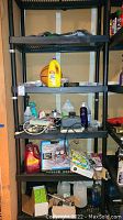 Full view of black plastic 4-tier shelf with assorted garage and cleaning supplies including Lysol cleaner, basketball, car wash liquid, batteries, light bulbs, tools and gauges.