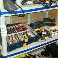 Shelf with Ryobi cordless drill, charger and multiple drill bit sets in plastic cases. Tools show dust and dirt with visible signs of rodent activity nearby.