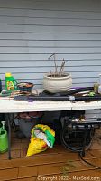 Shows a white folding table on a deck with multiple garden supplies including plant pots, garden soil bags, watering cans, and a pressure washer underneath.