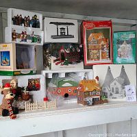 Wide view of the shelf containing multiple boxed and unboxed Christmas Village collectibles, including figurines, buildings, and animal accents.
