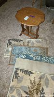 Wooden foldable end table on carpeted floor with seven small rugs in front of it, showing varying patterns.