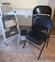 Photo showing two Cosco step stools, two Rubbermaid step stools, and three black padded folding chairs next to the folded table.