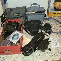 Photo showing a grey Bushnell branded fabric carrying bag, a grey fabric camera case, multiple black leather cases including a tripod bag, and an electronic item in a red box with a software CD.